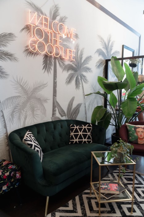 Grünes Samtsofa vor Palmentapete, darüber die Neonschrift „Welcome to the good life“